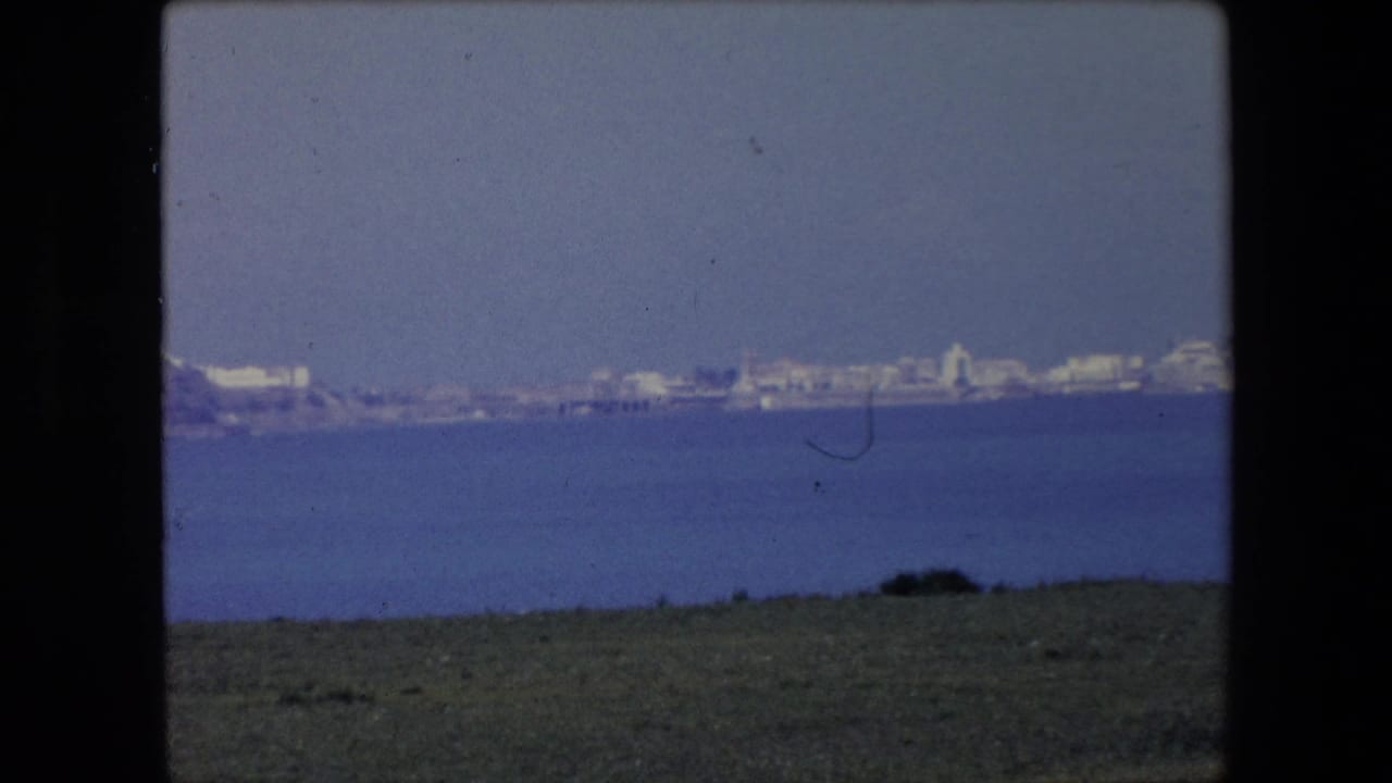 1971 Algeria 8mm frame with a wide distant shoreline, clustered pale buildings, blue water, and a narrow grassy foreground.