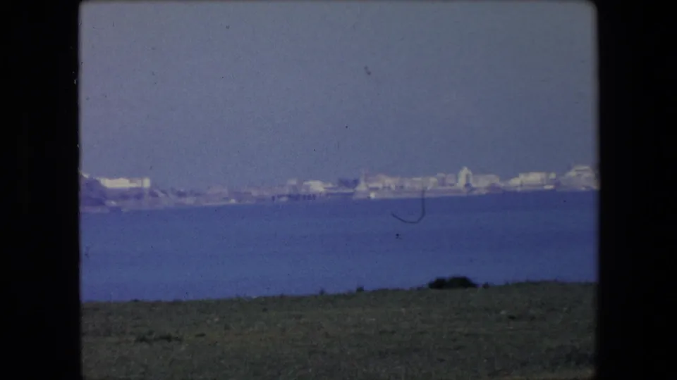 1971 Algeria 8mm frame with a wide distant shoreline, clustered pale buildings, blue water, and a narrow grassy foreground.