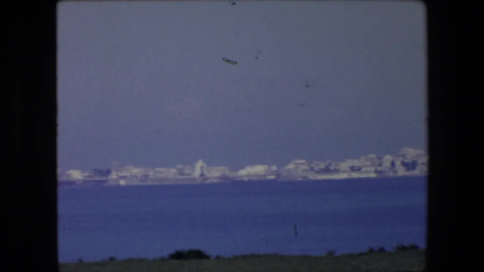 1971 Algeria home-movie frame showing a low coastal skyline of pale buildings across blue water, with grass along the lower edge and visible film grain.