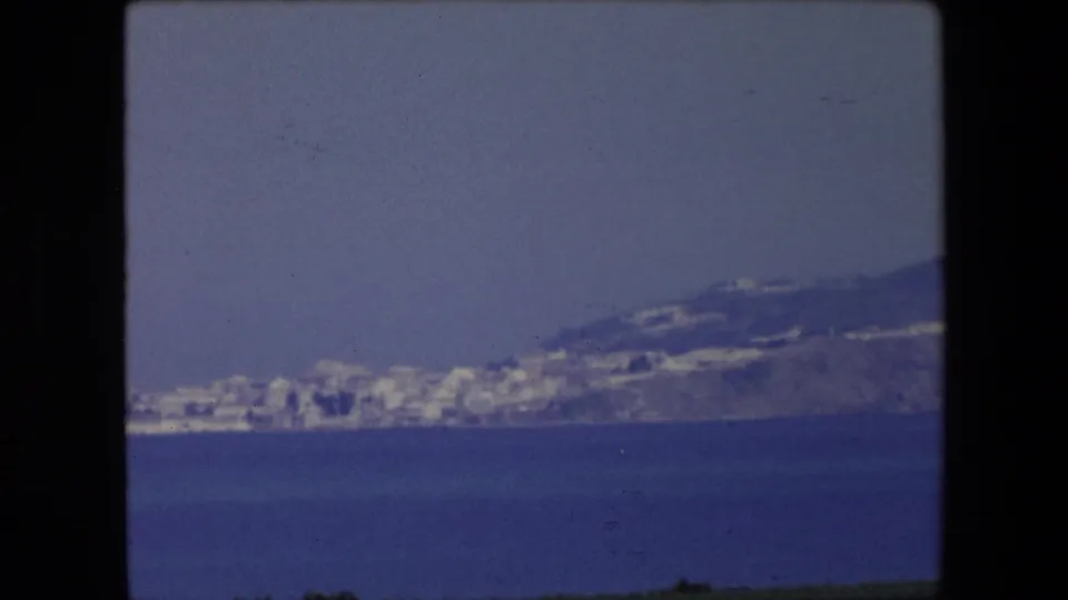 1971 Algeria 8mm frame showing white buildings along a distant hillside coast, with calm blue water in front and a soft hazy sky above.