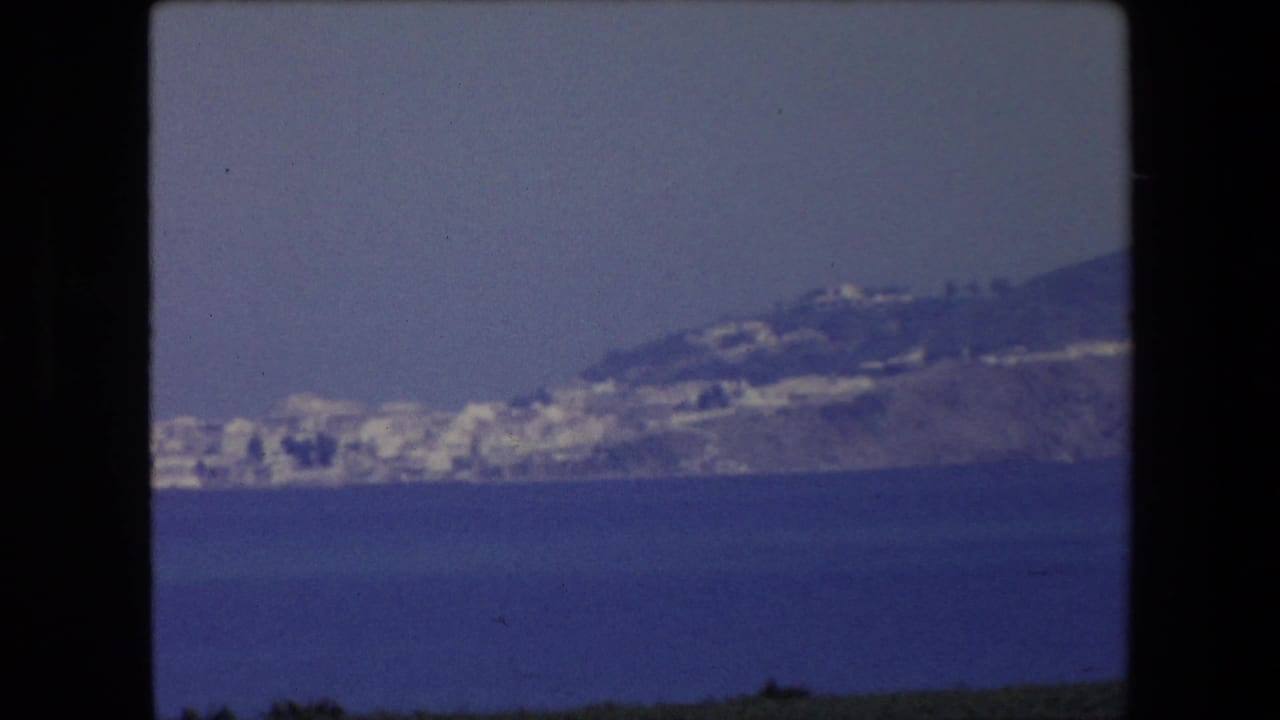 1971 Algeria home-movie frame showing a hazy coastal peninsula across the sea, with small white buildings clustered along the shoreline and hillside.