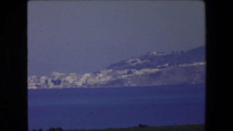 1971 Algeria home-movie frame showing a hazy coastal peninsula across the sea, with small white buildings clustered along the shoreline and hillside.
