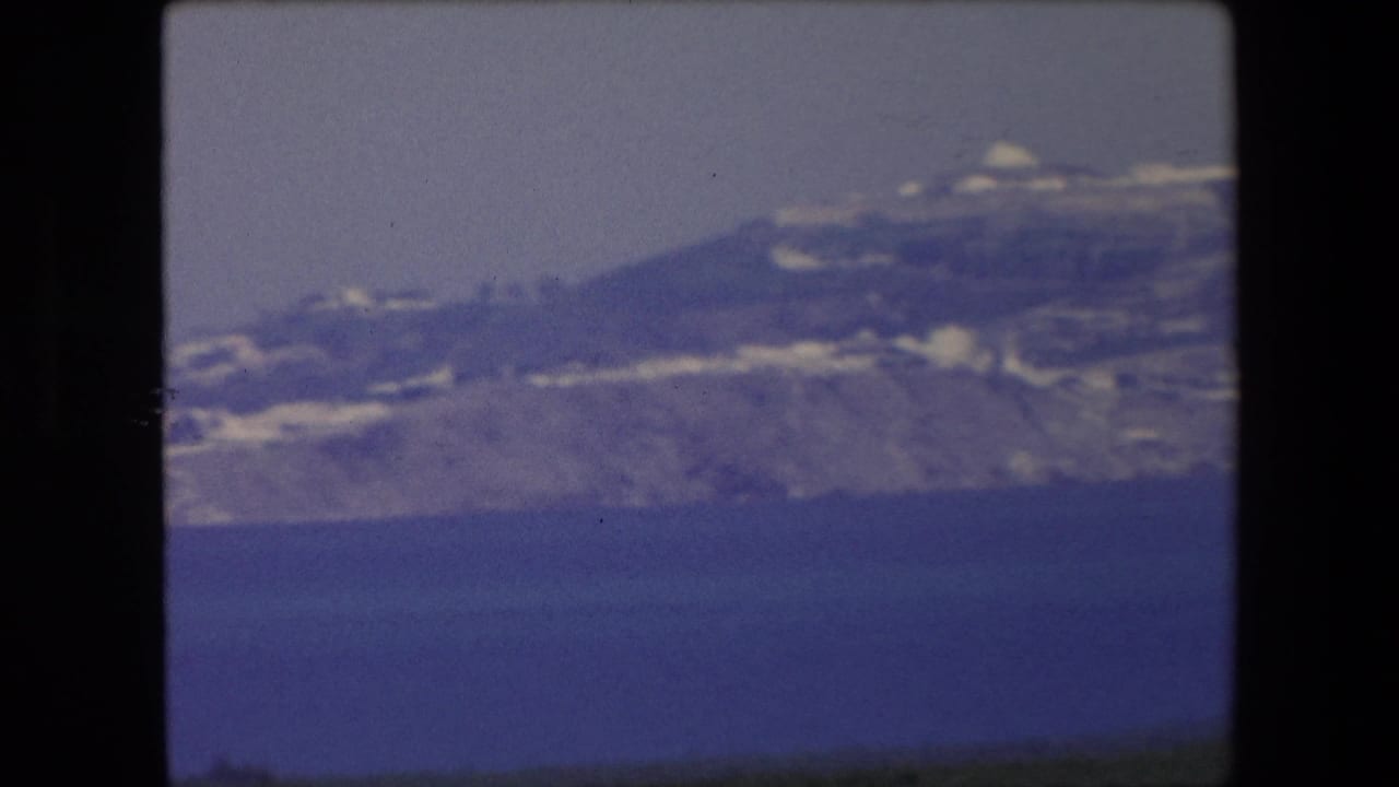 Closer soft view of a coastal hill with white structures above blue water.