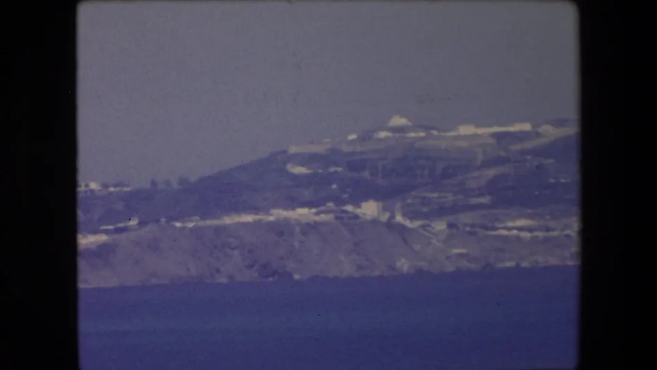 1971 Algeria frame showing a distant coastal hillside with white buildings, pale shoreline detail, and blue sea in the foreground.