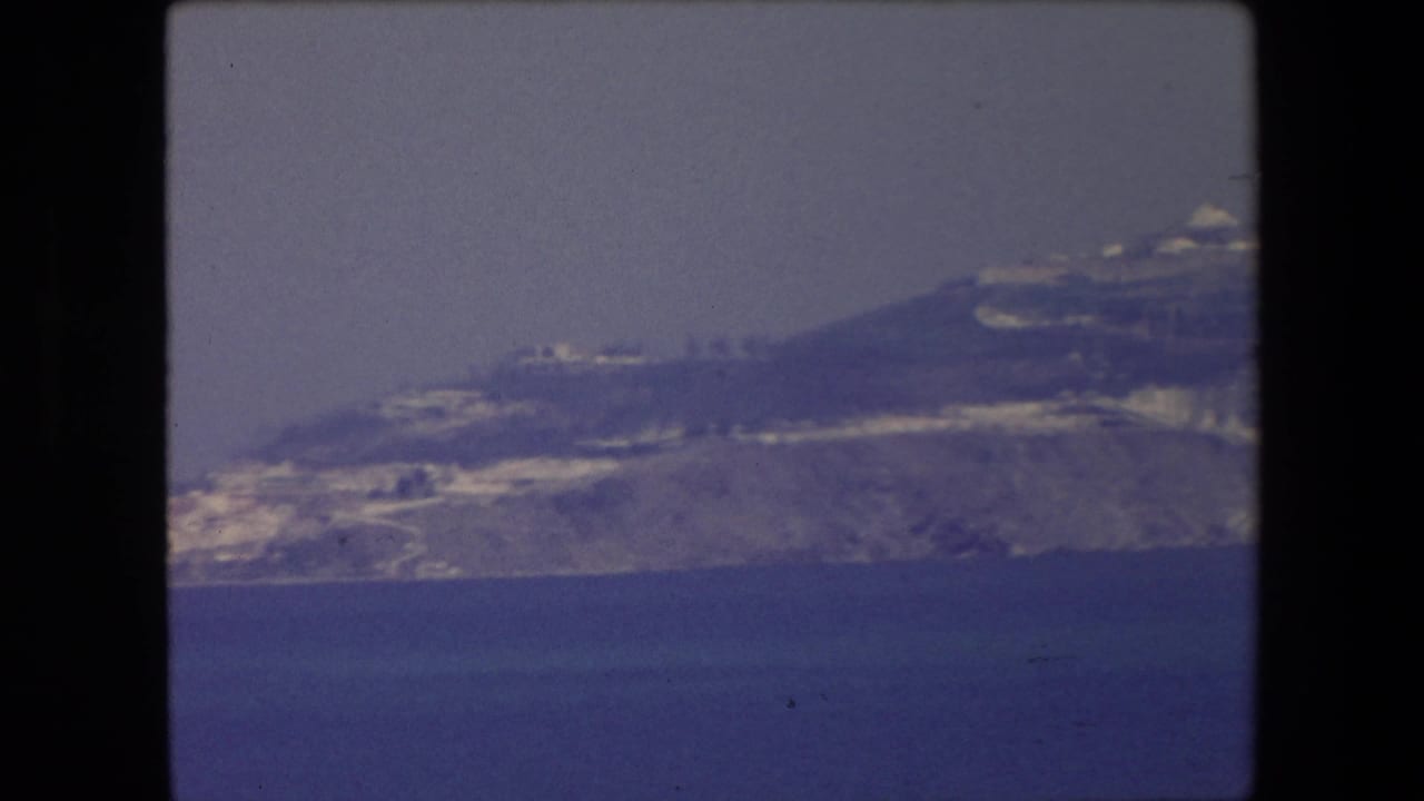 1971 Algeria 8mm frame showing a distant coastal headland or peninsula with pale buildings along the slope above deep blue water.