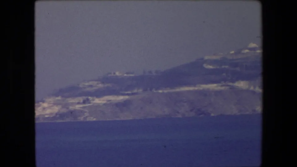 1971 Algeria 8mm frame showing a distant coastal headland or peninsula with pale buildings along the slope above deep blue water.