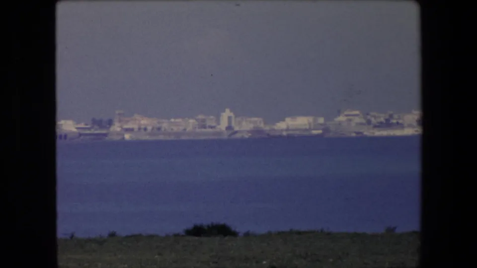 1971 Algeria 8mm frame showing a pale shoreline city across the water, with a strip of dark foreground land at the bottom of the view.