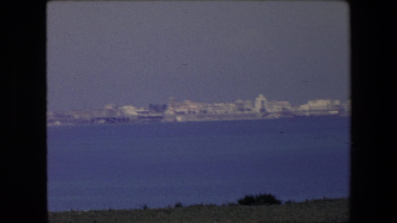 1971 Algeria 8mm frame showing a distant pale coastal city viewed across calm blue water, with a dark film border and soft haze.