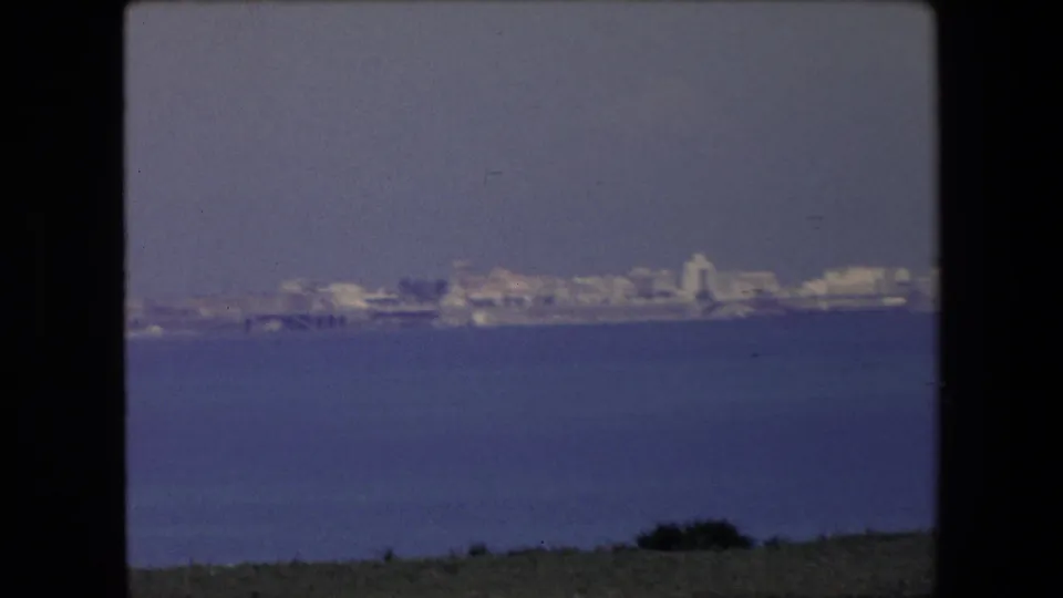 1971 Algeria 8mm frame showing a distant pale coastal city viewed across calm blue water, with a dark film border and soft haze.