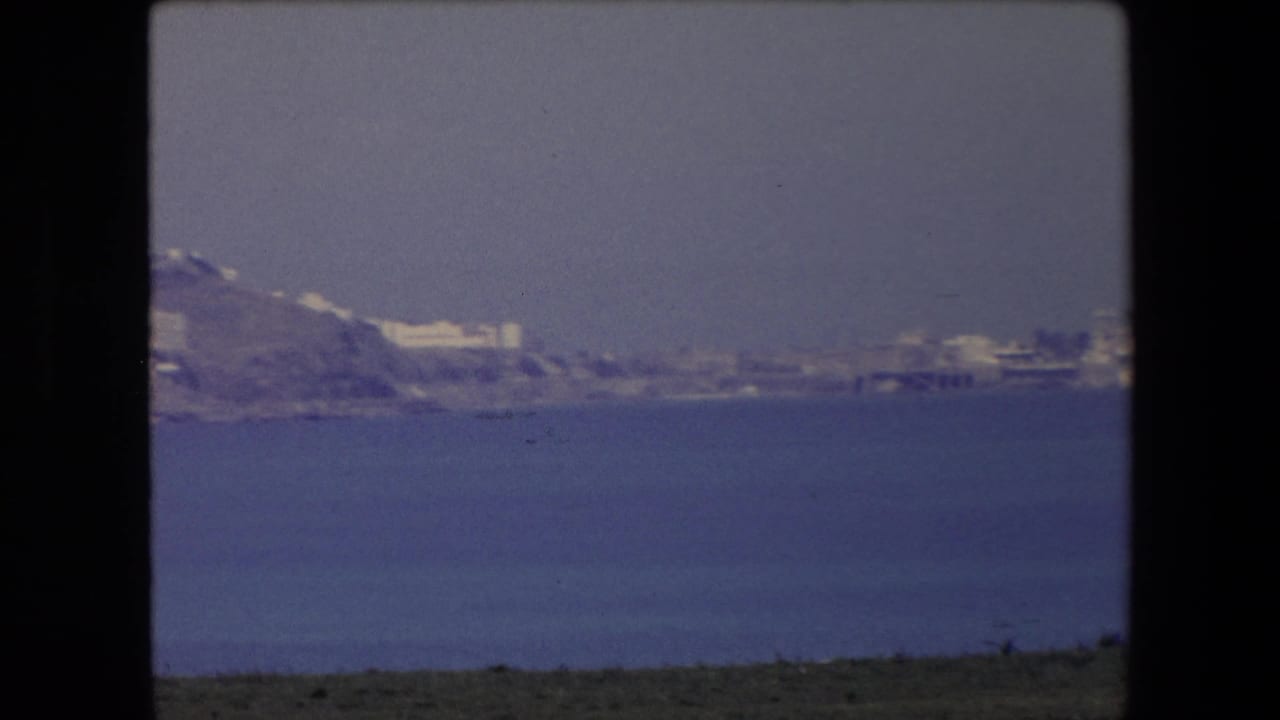 1971 Algeria home-movie frame with a far hillside city and shoreline seen from offshore across a wide band of blue sea.