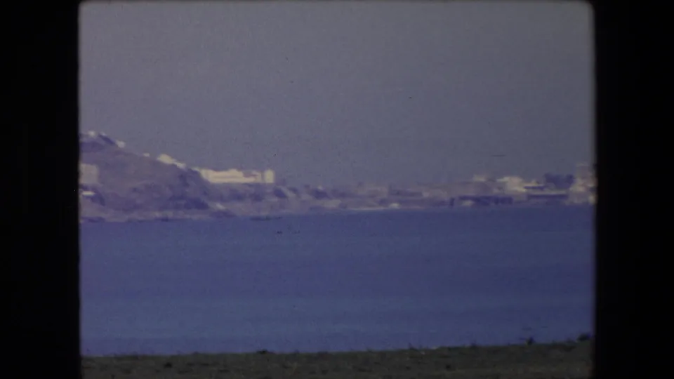 1971 Algeria home-movie frame with a far hillside city and shoreline seen from offshore across a wide band of blue sea.