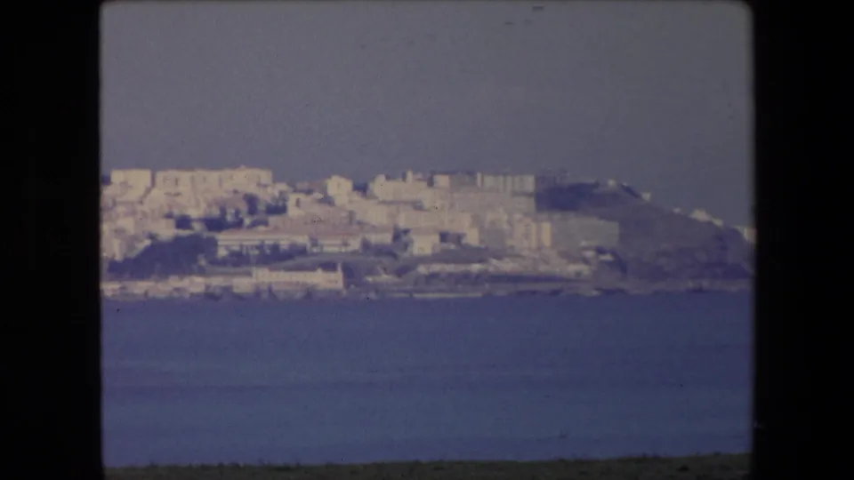1971 Algeria frame showing a distant coastal hillside city with pale buildings, calm sea in the foreground, and a hazy sky.