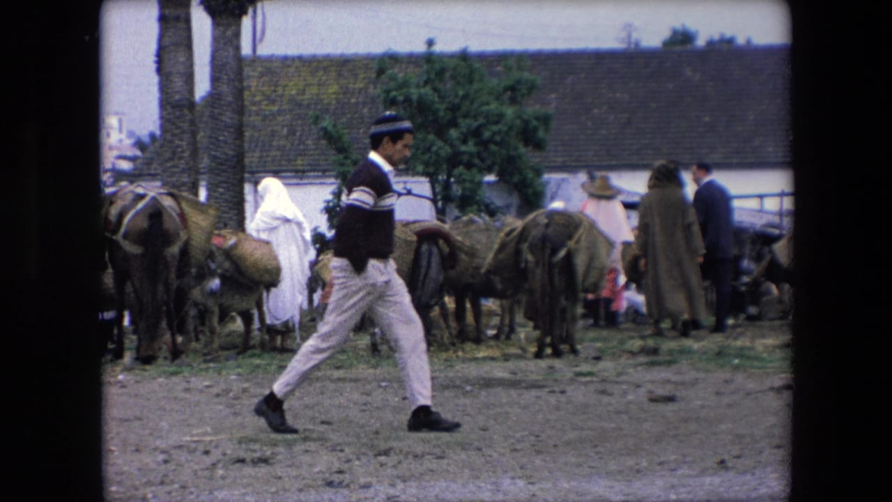In 1971 Algeria, a man in a striped sweater and cap walks across the foreground while people and loaded donkeys gather near low buildings and palm trees.