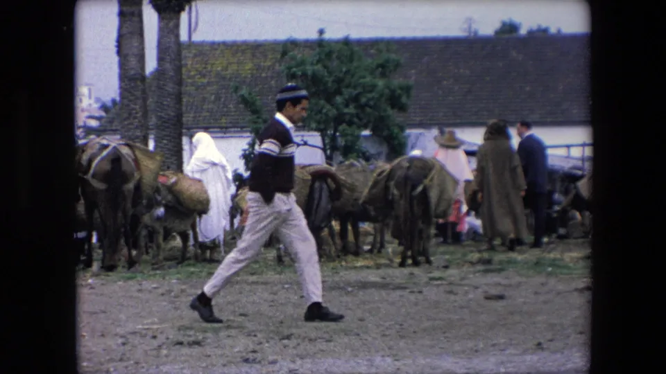 In 1971 Algeria, a man in a striped sweater and cap walks across the foreground while people and loaded donkeys gather near low buildings and palm trees.