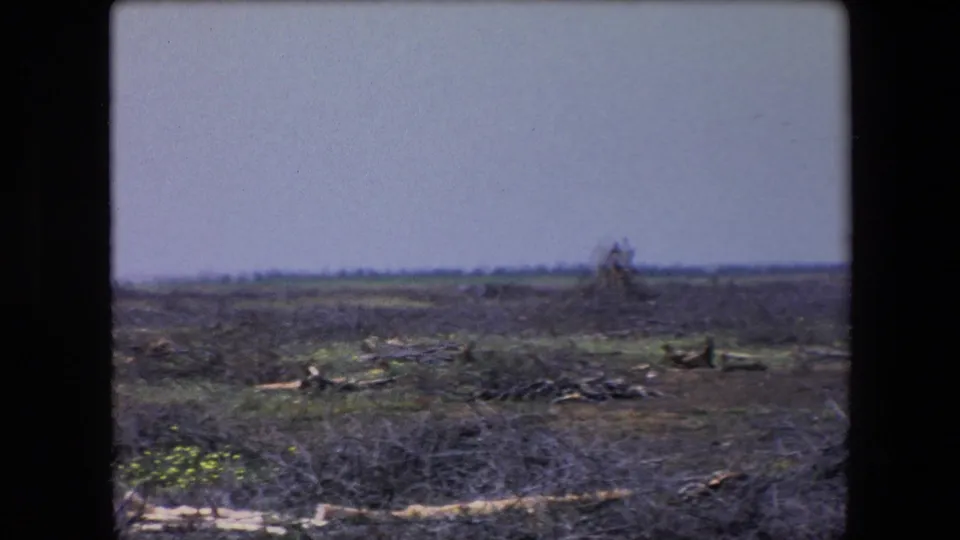 1971 Algeria 8mm frame showing a wide, barren field with scrub, debris, and a small collapsed form near the horizon.