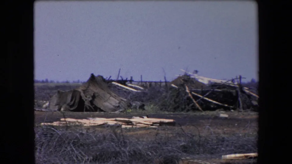 1971 Algeria 8mm frame with tattered canvas, leaning wooden poles, scattered planks, sparse vegetation, and a broad pale sky.