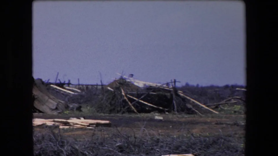1971 Algeria 8mm frame showing collapsed fabric at the left edge, broken pole framework, loose boards, scrub, dark soil, and distant flat terrain.