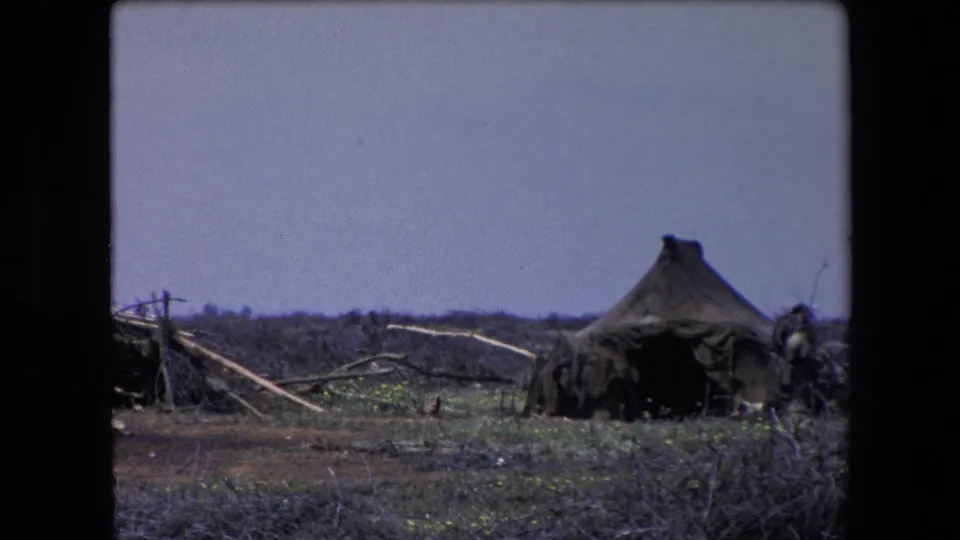 1971 Algeria 8mm frame showing an upright dark canvas form beside fallen poles, scrub vegetation, dark ground, and a low distant horizon.