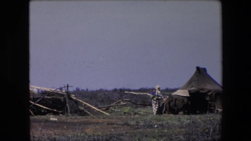 1971 Algeria 8mm frame with a distant person in patterned clothing standing or walking near a dark shelter beside a conical thatched hut.