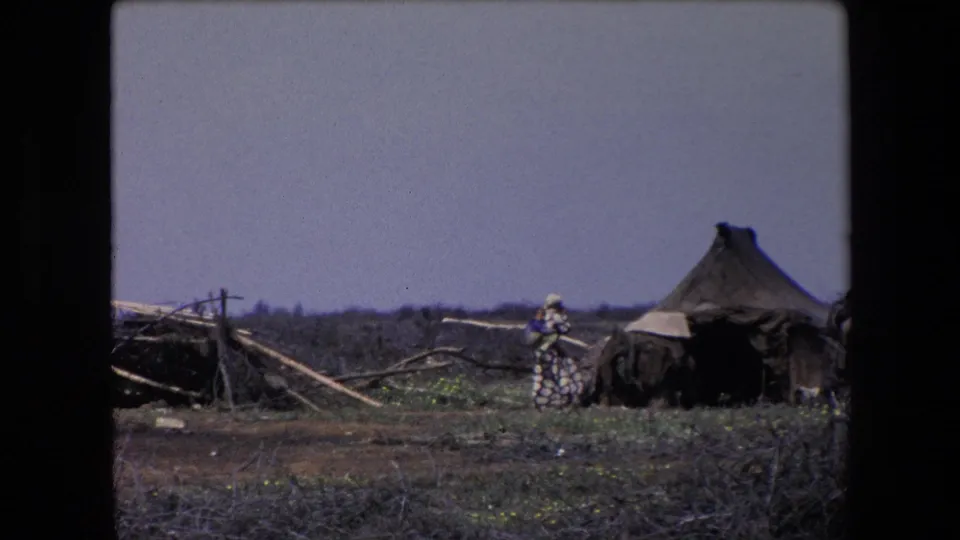 1971 Algeria 8mm frame showing a distant figure near a dark tarp-covered shelter, with a conical thatched hut and scattered branches in the background.