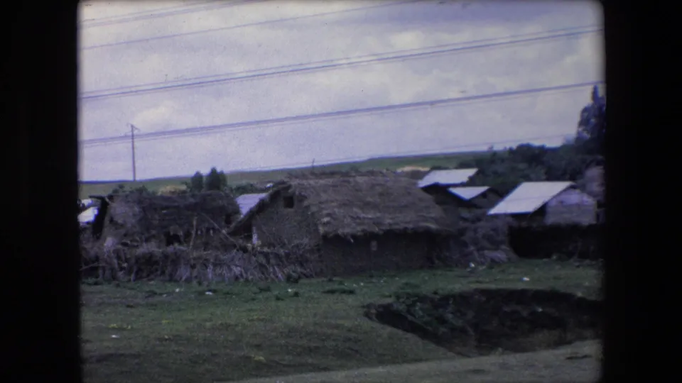 A wider rural view shows low thatched-roof structures and grassy ground below a road, with the 8mm frame split by dark film borders.