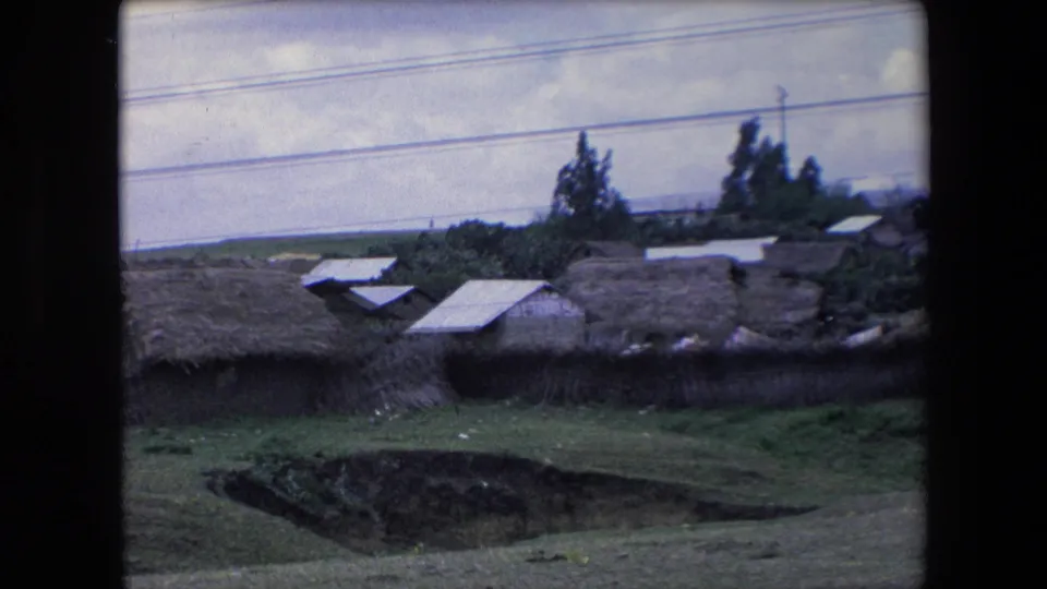 1971 Algeria home-movie frame showing thatch-roofed village structures beyond grassland, with trees, cloudy sky, and utility lines overhead.