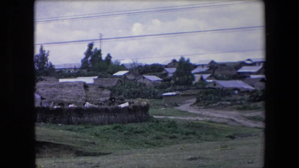 1971 Algeria home-movie frame showing a rural settlement with thatched buildings, pale roof panels, shrubs, a dirt road, and utility lines across the sky.