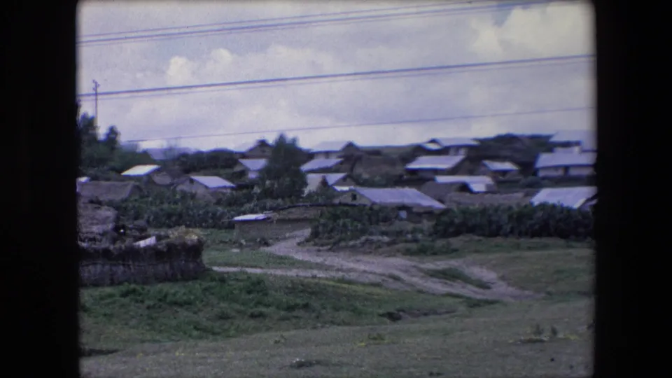 1971 Algeria home-movie frame with a wider view of low village buildings, a winding dirt road, grassy open ground, trees, cloudy sky, and overhead lines.
