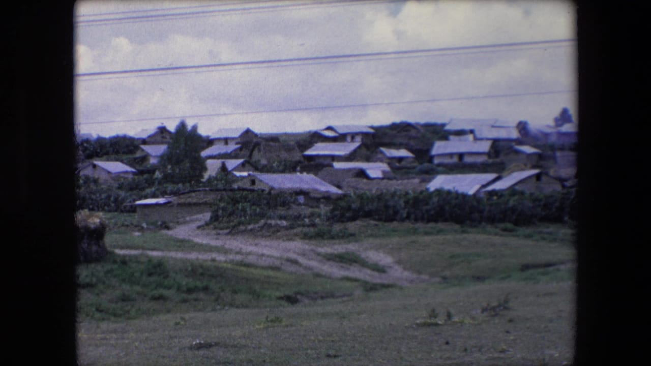 Wide view of a rural hillside village with low roofs, fields, dirt tracks, and power lines.