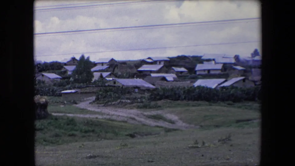 1971 Algeria 8mm frame showing a wide rural hillside village with modest homes, green open ground, dirt paths, and overhead utility lines beneath a cloudy sky.