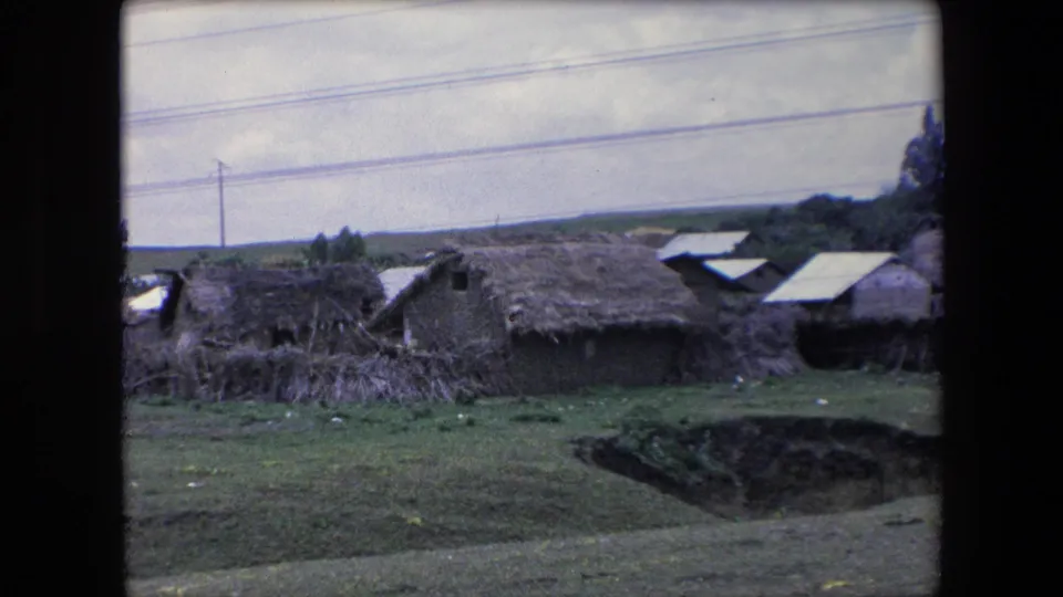 1971 Algeria 8mm frame moving closer to rural dwellings, with thatched hut roofs, corrugated roof panels, open grass, utility poles, and a dark ditch in the foreground.