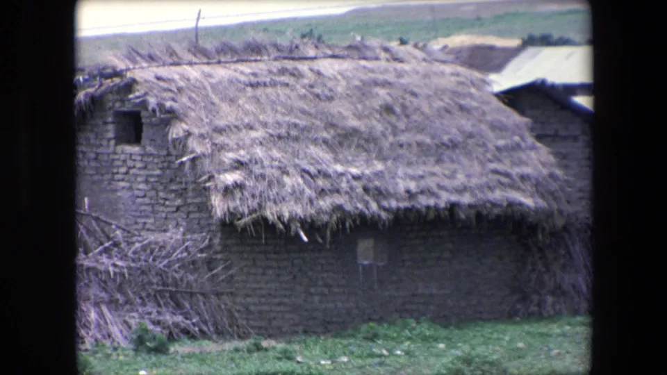 The frame shows a weathered thatched dwelling in Algeria, filmed on 8mm in 1971, with a small window, brush, grass, and low open terrain behind it.