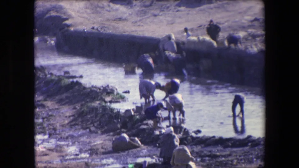1971 Algeria 8mm frame showing people gathered along a shallow water channel, with figures bending over laundry or fabric at the edge.