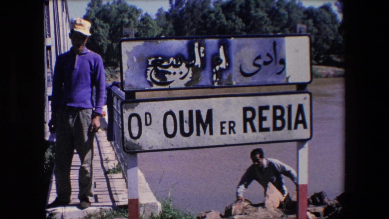 A man in a hat stands beside a weathered Arabic and Latin Oum Er-Rbia sign near a bridge, with another man visible by rocks below.