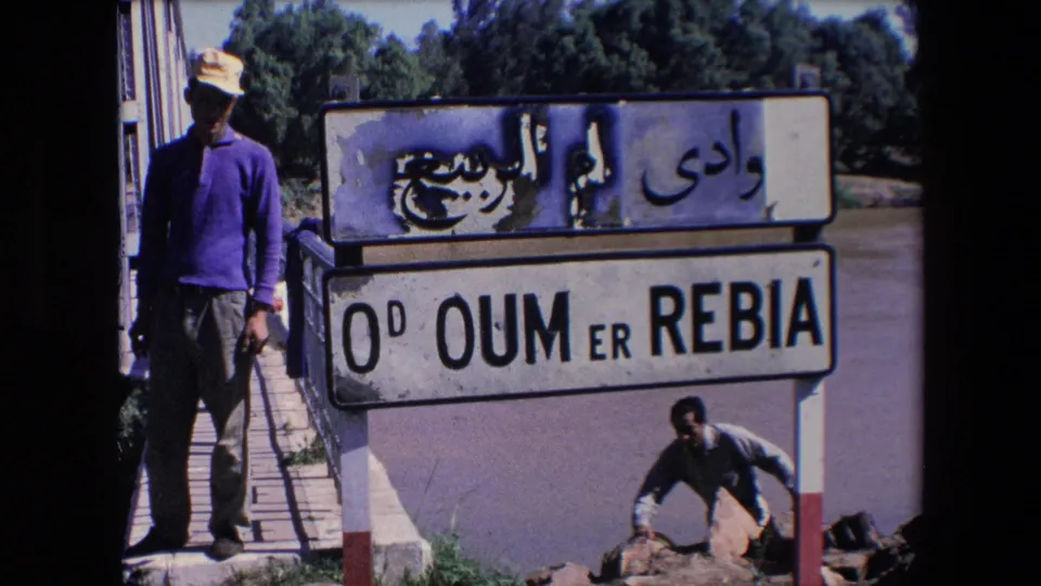 A man in a hat stands beside a weathered Arabic and Latin Oum Er-Rbia sign near a bridge, with another man visible by rocks below.