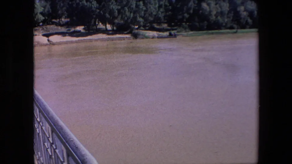 A broad muddy river extends past a bridge railing toward a tree-lined bank in faded early-1970s 8mm travel footage.