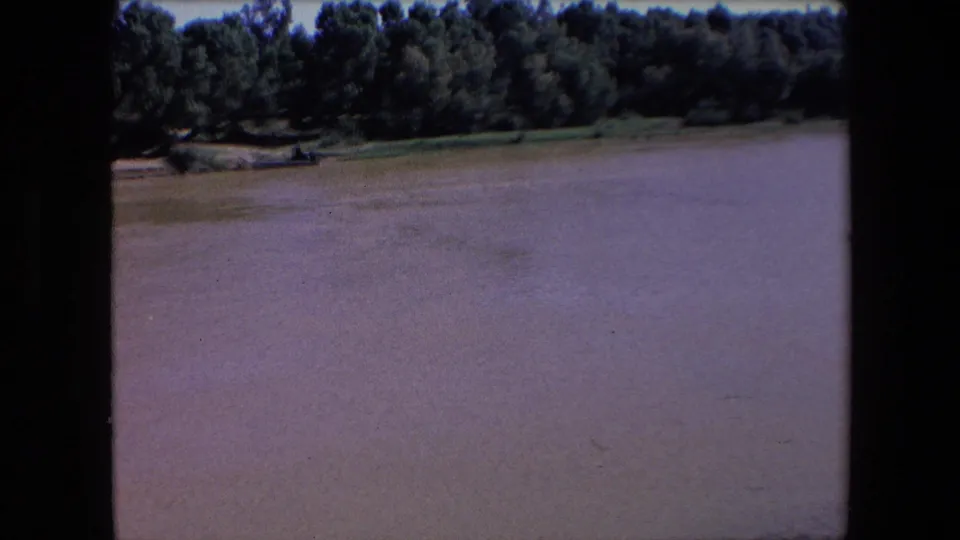 1971 Algeria 8mm frame showing a wide muddy water surface under a dark tree line, with the far shore running horizontally across the upper portion of the view.