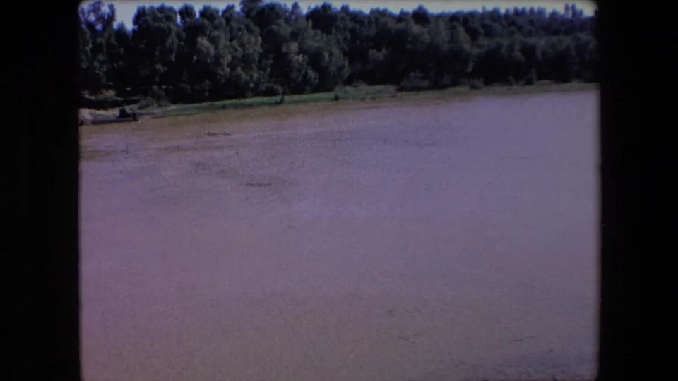1971 Algeria 8mm frame of the same quiet muddy river, with a dense wooded far bank and visible film-frame borders emphasizing the scanned home-movie source.