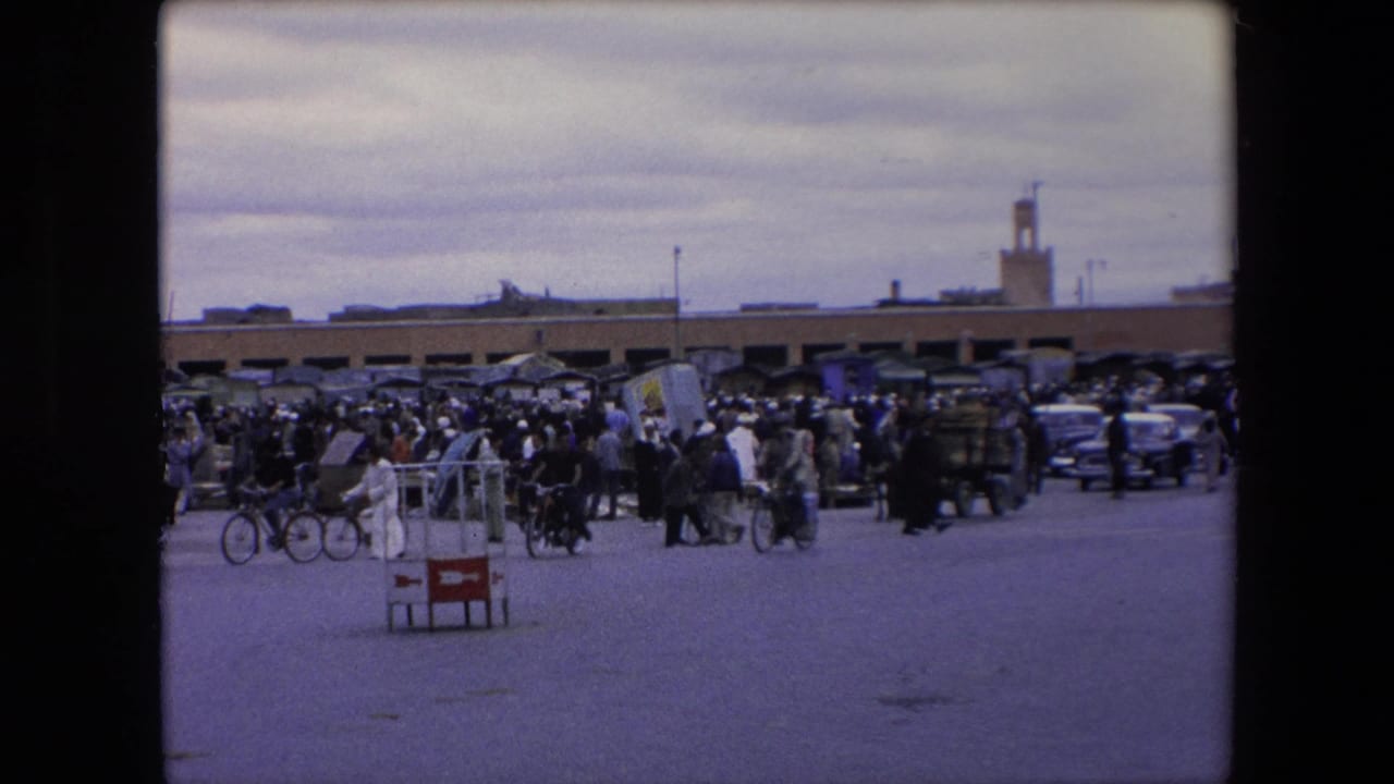 Crowded street market with people, bicycles, cars, carts, and a minaret in the distance.