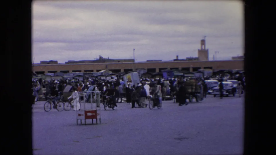 1971 Algeria 8mm frame showing a busy public market street with pedestrians, bicycles, parked cars, carts, stalls, and a minaret beyond low buildings.