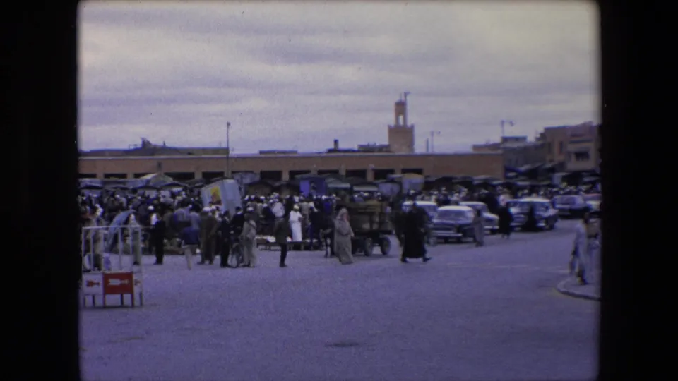 1971 Algeria street-market frame with people gathering near carts and stalls, cars along the roadway, and a minaret visible in the background under an overcast sky.