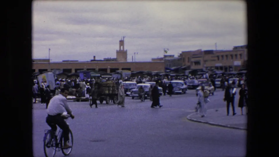 1971 Algeria 8mm frame with a bicyclist in the foreground, people crossing the street, carts and cars near a market, and a minaret beyond the low roofline.