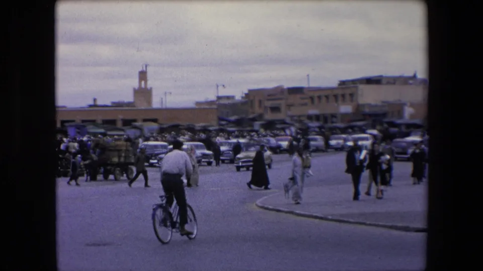 1971 Algeria street scene showing a bicyclist riding away from the camera, pedestrians walking near market carts, cars lined across the street, and a minaret in the background.