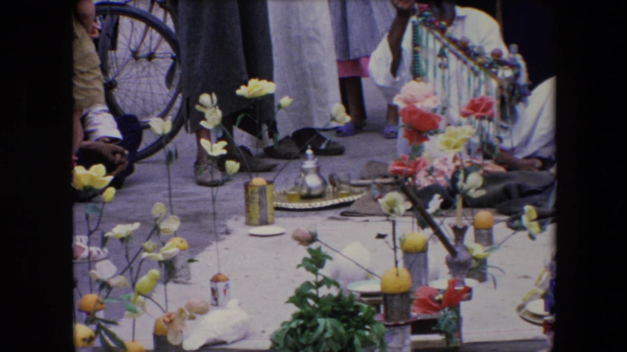1971 Algeria 8mm frame showing an outdoor religious-event display with artificial flowers, lemons, a metal teapot on a tray, seated and standing people, and a bicycle wheel at the edge of the scene.