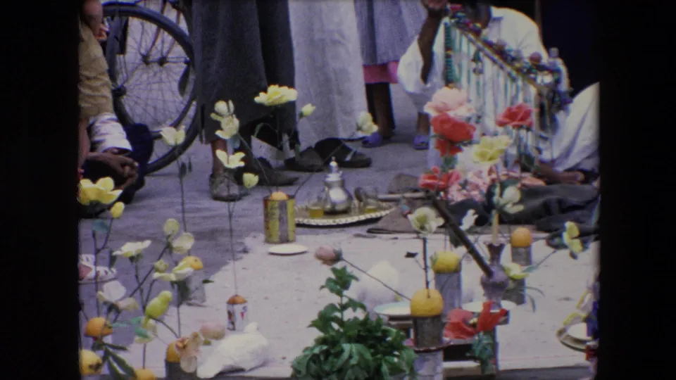 1971 Algeria 8mm frame showing an outdoor religious-event display with artificial flowers, lemons, a metal teapot on a tray, seated and standing people, and a bicycle wheel at the edge of the scene.