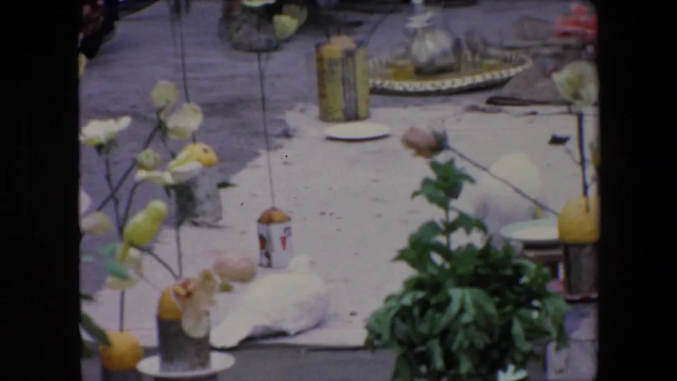 1971 Algeria 8mm frame centered on a pavement display with flowers in jars, lemons or yellow fruit, small containers, a white cloth bundle, and a leafy plant during an outdoor religious event.