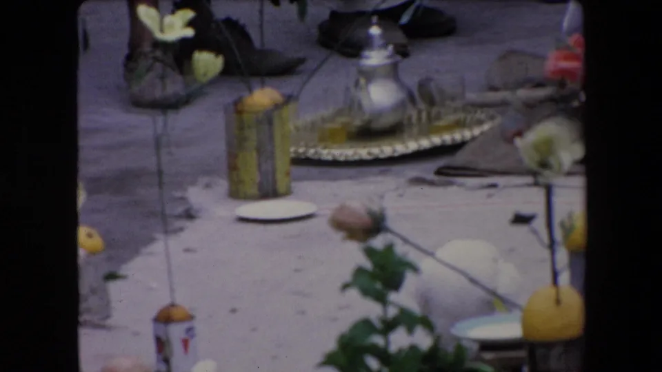 1971 Algeria 8mm frame showing a metal tray with a silver teapot, nearby flowers, lemons or yellow fruit, small containers, shoes, and other display objects at an outdoor religious gathering.