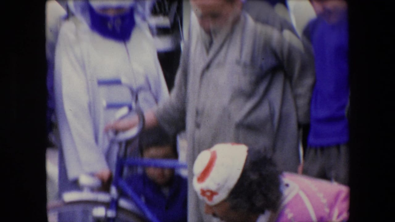 1971 Algeria home-movie frame showing an outdoor crowd with standing onlookers and a partially visible seated figure in pink and white clothing near the lower edge.