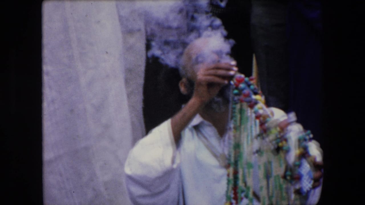 Man in white clothing smoking beside colorful hanging beads with onlookers behind him.