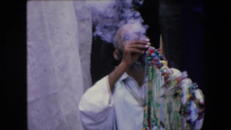1971 Algeria frame of a man in white clothing smoking while holding or sitting beside colorful hanging beads, with people standing behind him outdoors.